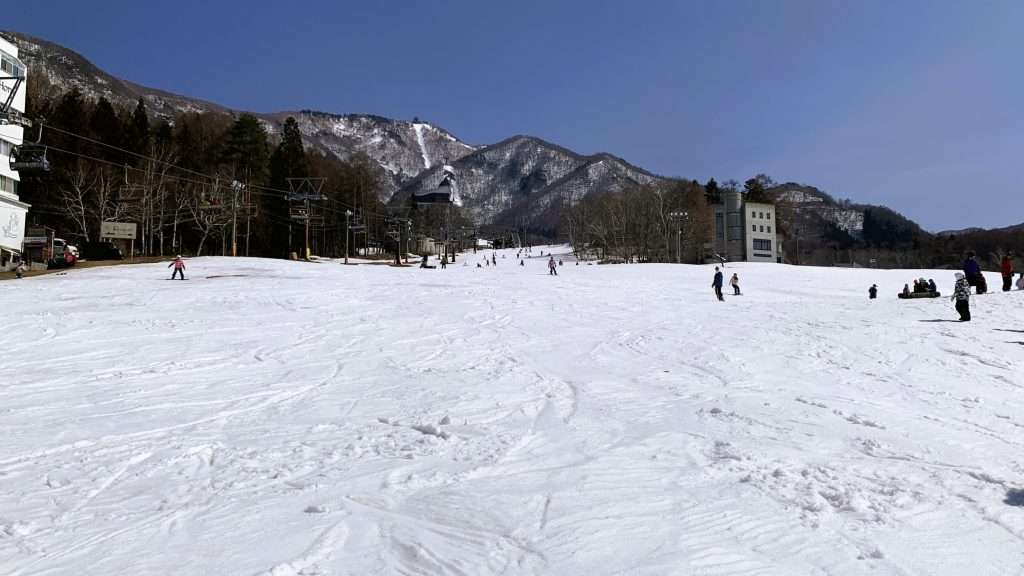 長野県北志賀竜王スキーパークBaseゲレンデを見上げる冬景色。一面に広がる雪のゲレンデにはスキーやスノーボードの滑走跡が残り、左手のリフトと程よく滑る人々の姿。奥には雪山と青空が広がる自然豊かなパノラマ。