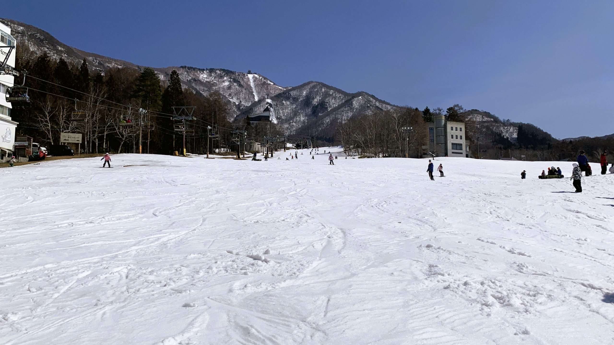 長野県北志賀竜王スキーパークBaseゲレンデを見上げる冬景色。一面に広がる雪のゲレンデにはスキーやスノーボードの滑走跡が残り、左手のリフトと程よく滑る人々の姿。奥には雪山と青空が広がる自然豊かなパノラマ。