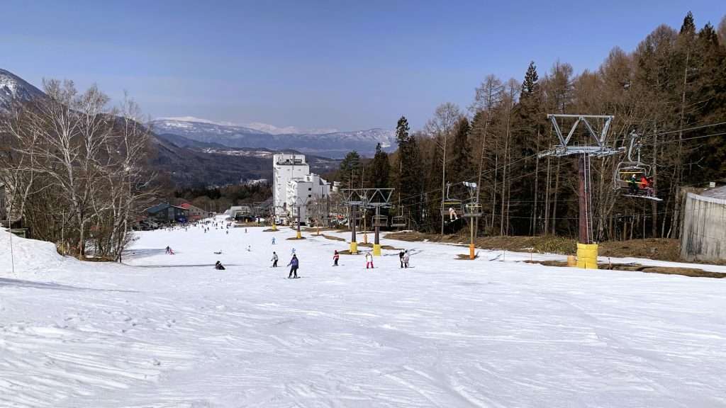 北志賀竜王スキーパークBaseゲレンデ上空からの俯瞰ビュー。雪に刻まれたスキー・スノーボード跡、リフトと滑るスキーヤー・ボーダー、木々に囲まれたゲレンデ、遠くに見える山々と青空が広がる晴天の雪景色。