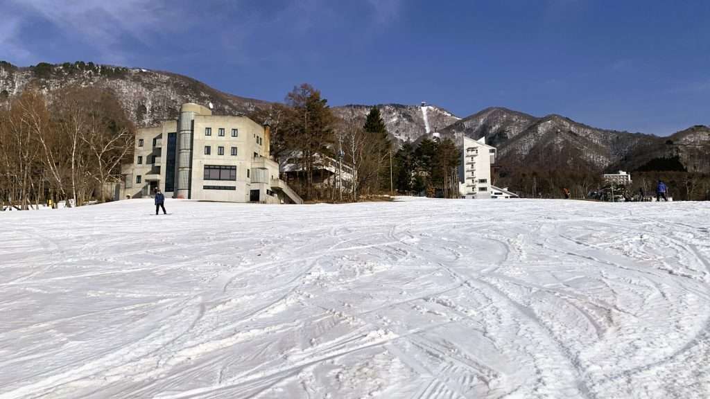 北志賀竜王スキーパークのゲレンデを見上げる形で撮影。雪に残るスキーやスノーボードの滑走跡、ほとんど人の姿がない静かな雰囲気。奥に見える木々や建物、山々と青空が広がる晴天の冬景色。