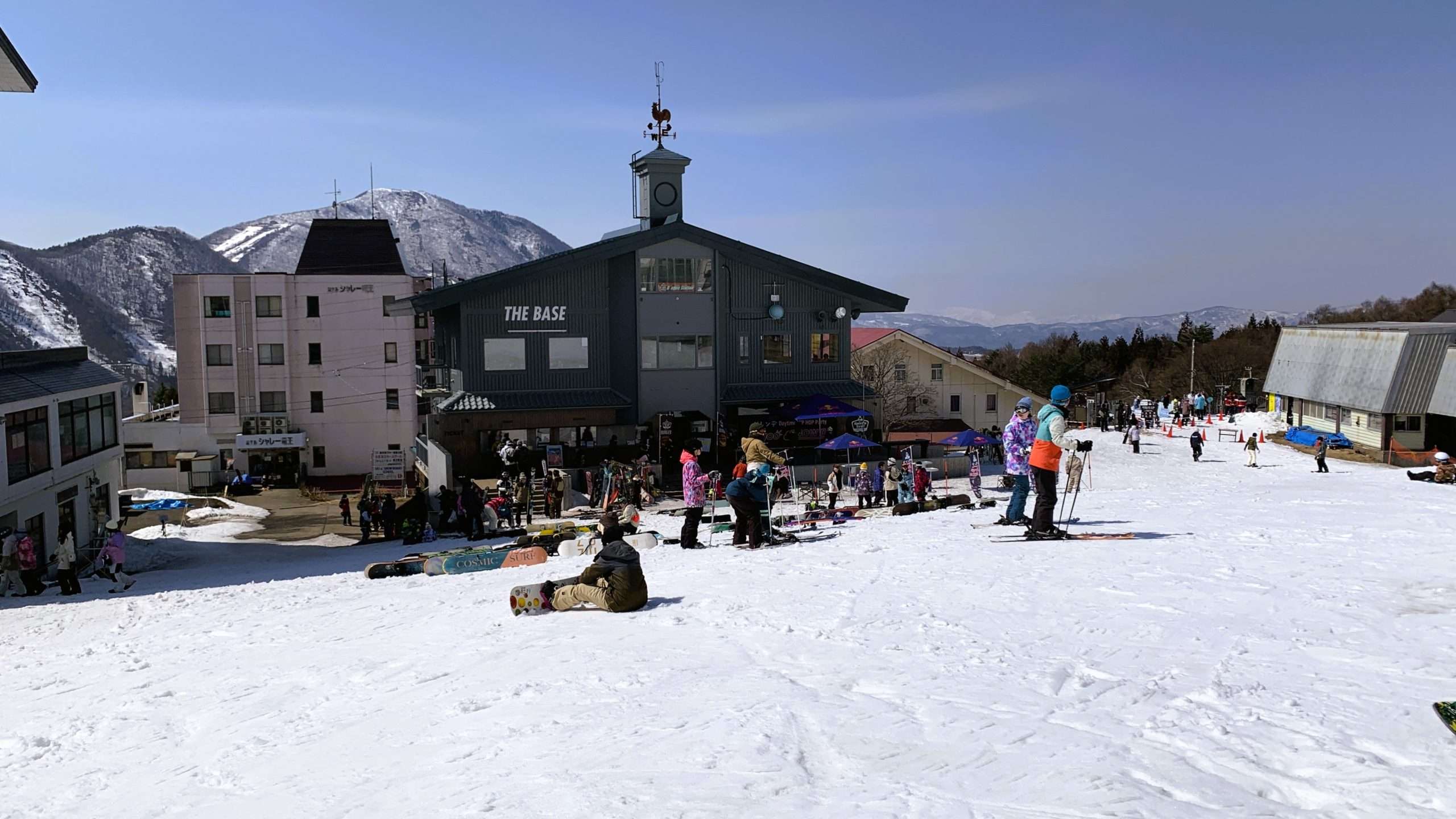 長野県北志賀竜王スキーパークTHE BASE付近を撮影。風見鶏がある黒い三角屋根の建物。多くのスノーボーダーやスキーヤー、置かれたスノーボード、周囲のホテル建物、奥に見える山々、晴れた日の冬のゲレンデ風景。
