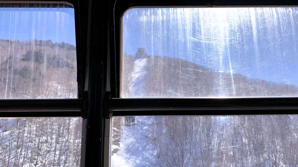 北志賀竜王スキーパークのロープウェイ内部からの視点。山頂へ向かう雪山を見上げる構図。窓フレームの影と白い雪景色のコントラスト。頂上駅と青空が映える晴れた日の山岳風景。