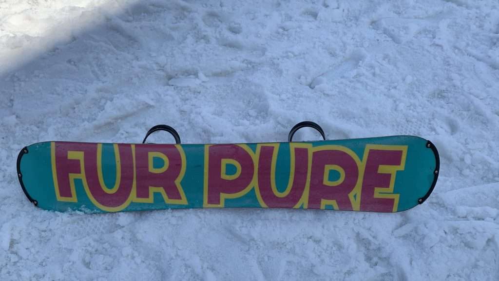 スノーボードの板の裏面デザインを雪のゲレンデ上で撮影。青緑のベースに「FOR PURE」のピンク文字が大きく入ったグラフィック。冬のスノーボードシーンと用具のディテールがわかる写真。