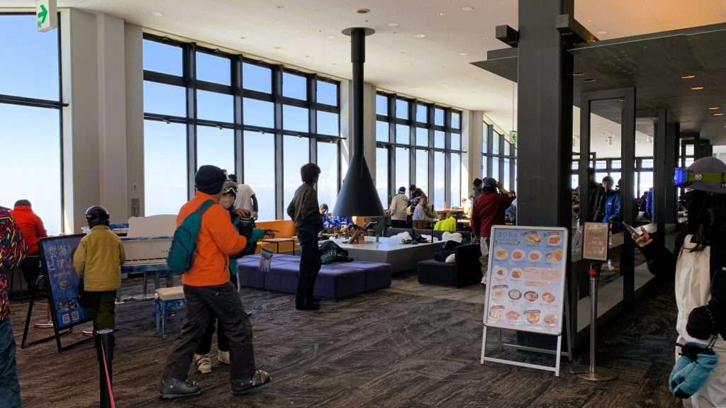 長野県北志賀竜王スキーパーク、ロープウェイ山頂付近のSORA terrace cafe店内。黒いじゅうたん、白いピアノ、暖炉などおしゃれなインテリア。多くの人で賑わい、奥の窓から青空が見える冬の山頂カフェ風景。