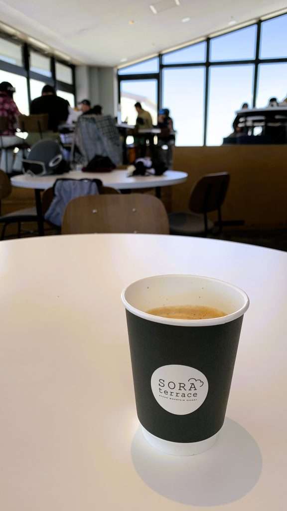 SORA terrace cafeで提供されるカフェラテ。紙コップに入ったロゴ付きドリンクをテーブル上で撮影。背景に店内と来店客、窓越しに青空が見える山頂カフェのリラックスした雰囲気。