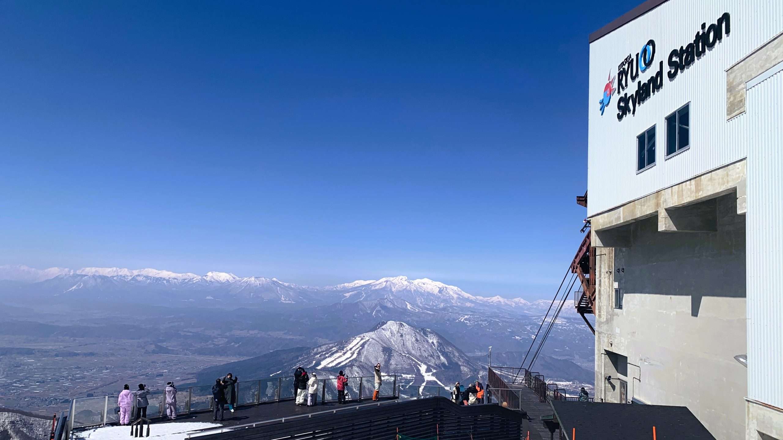 長野県北志賀竜王スキーパーク、ロープウェイ山頂付近のSORA terraceを撮影。右手にロープウェイ建物、手前に展望台があり程よく人が写真撮影。青空の下に広がるアルプス山脈と晴天の山頂景色。