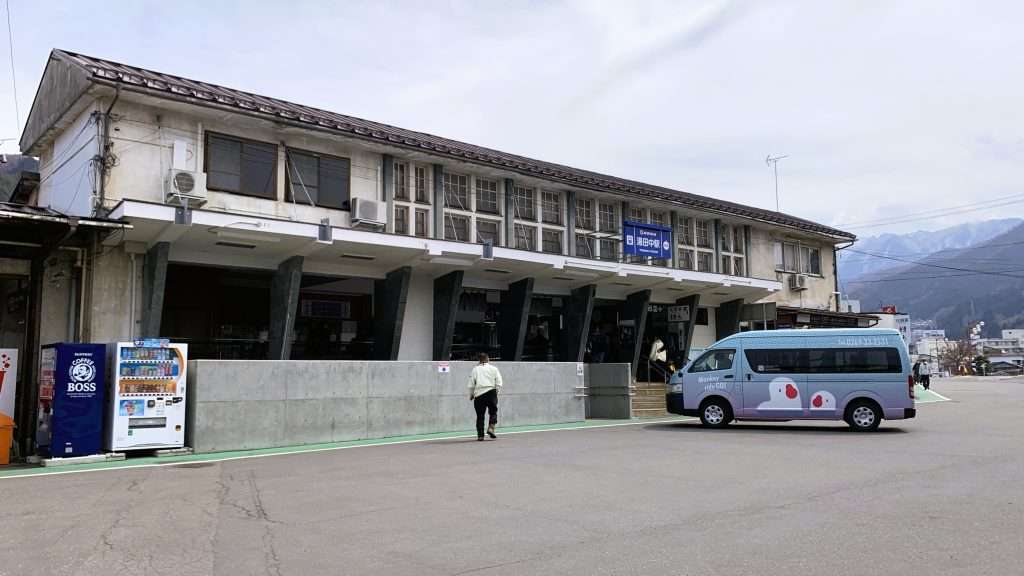長野県湯田中にある長野電鉄湯田中駅の駅舎を撮影。2階建ての歴史を感じる外観。温泉地へのアクセス拠点として利用される駅と周辺の街並み。曇り空が広がるローカルな風景。