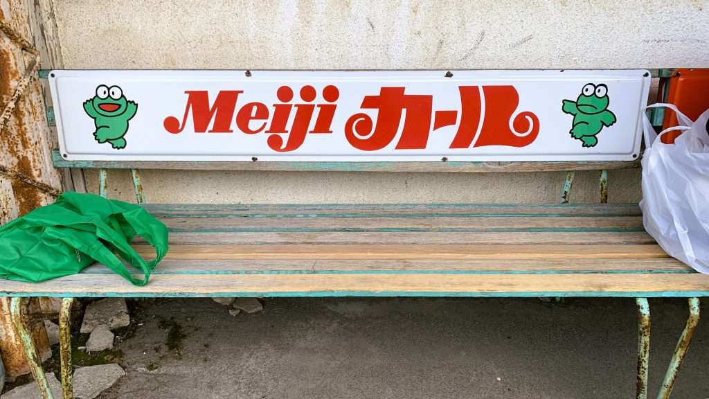 湯田中駅前、木製で塗装が剥げた昭和レトロの緑色のベンチ。背もたれには、meijiカールの看板。かわいいカエルのイラスト付き。駅前に残るレトロな街並みの一コマを捉えた写真。