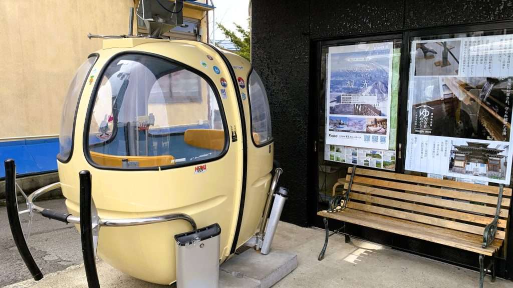 長野県湯田中駅前、観光案内所前にある丸型ロープウェイを撮影。クリーム色の丸みあるフォルムに4つの窓。右側に黒壁の観光案内所、前に木製ベンチが置かれた風景。