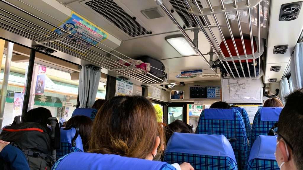 奈良交通やまかぜ号の車内を撮影。新宮駅方面へ向かう満席のバス、リュックを抱えた利用客、青いチェック柄シートと窓のカーテン、晴天の車窓が見える移動風景。