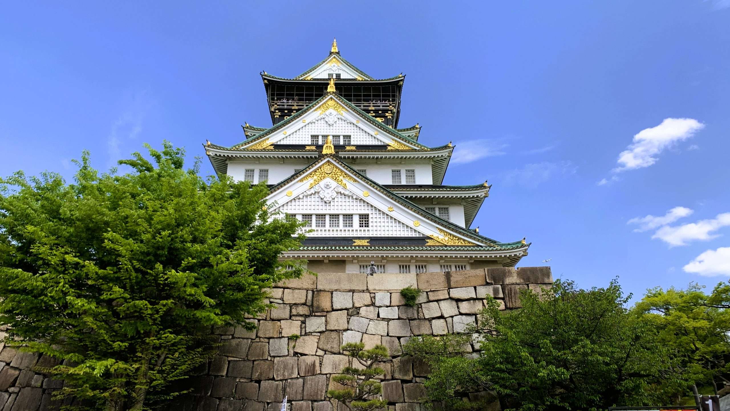 大阪城を正面から撮影した風景。石垣の上にそびえる白壁と金色の装飾が特徴的な天守閣、周囲を囲む緑の木々と青空の下で映える大阪の代表的な観光名所。