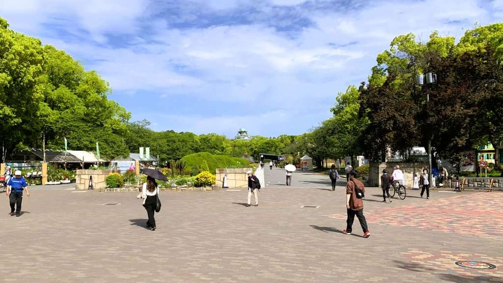 大阪城公園内の森ノ宮駅側入口を撮影した風景。石のタイルが一面に敷かれた広場と周囲を囲む緑の樹木、奥に小さく見える大阪城天守閣、歩く人々と青空にうっすら白い雲が浮かぶ大阪の観光入口の景観。