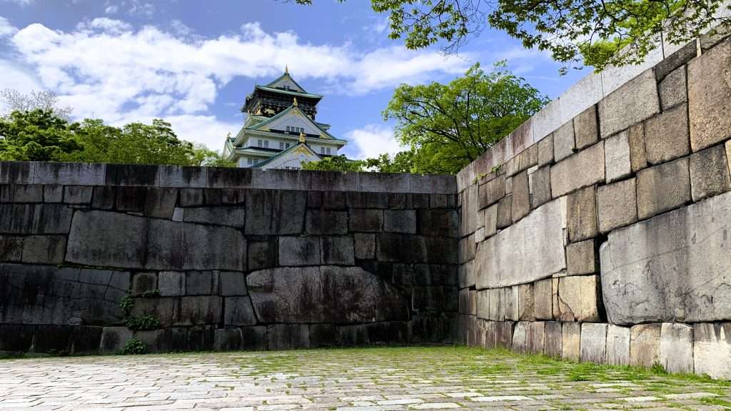 大阪城公園内の石垣エリアを撮影した風景。正面と右側に続く巨大な石垣と足元の石畳、奥に大阪城天守閣が見え、周囲に緑の木々と青空と白い雲が広がる大阪の代表的観光スポットの景観。