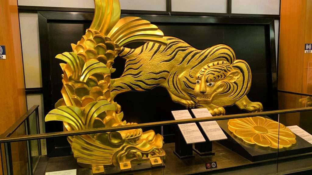 大阪城天守閣2階の展示スペースを撮影。金のしゃちほこと虎や菊の装飾モチーフが並ぶ館内展示、背景には黒い壁面パネルが使われた観光展示エリアの風景。