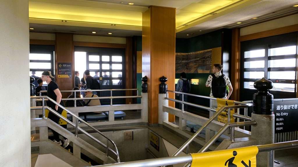 大阪城天守閣の最上階展望フロアを撮影した館内風景。中央に階段がありその周囲を回遊できる通路が設けられ、外へ出られる扉が複数並ぶ構造。木材と石材が使われた空間に観光客の姿が見える大阪観光スポットの様子。