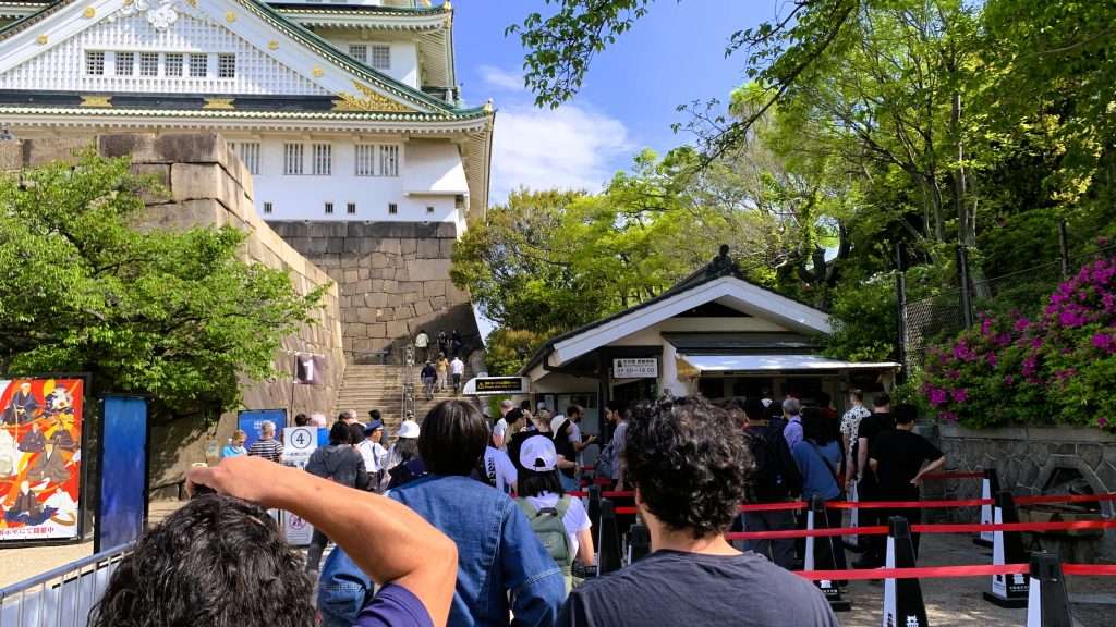 大阪城天守閣のチケット売り場前の列を撮影した風景。赤い誘導レーンで区切られた導線に観光客が並ぶ様子、右奥に屋根付きチケット売り場、左側に大阪城の石垣と下部構造、緑の木々と晴天が広がる観光地の一場面。