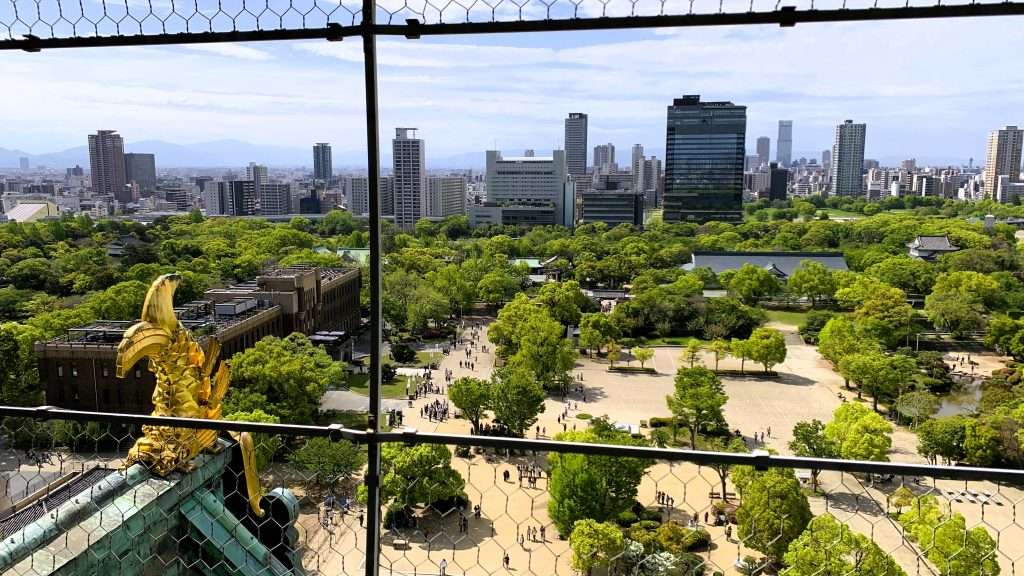 大阪城天守閣最上階の展望フロアから撮影した風景。左手前に金のしゃちほこが見え、眼下には緑豊かな大阪城公園と歩く人々、奥に広がる大阪市街のビル群と青空に浮かぶ雲が見える都市と歴史が融合した景観。