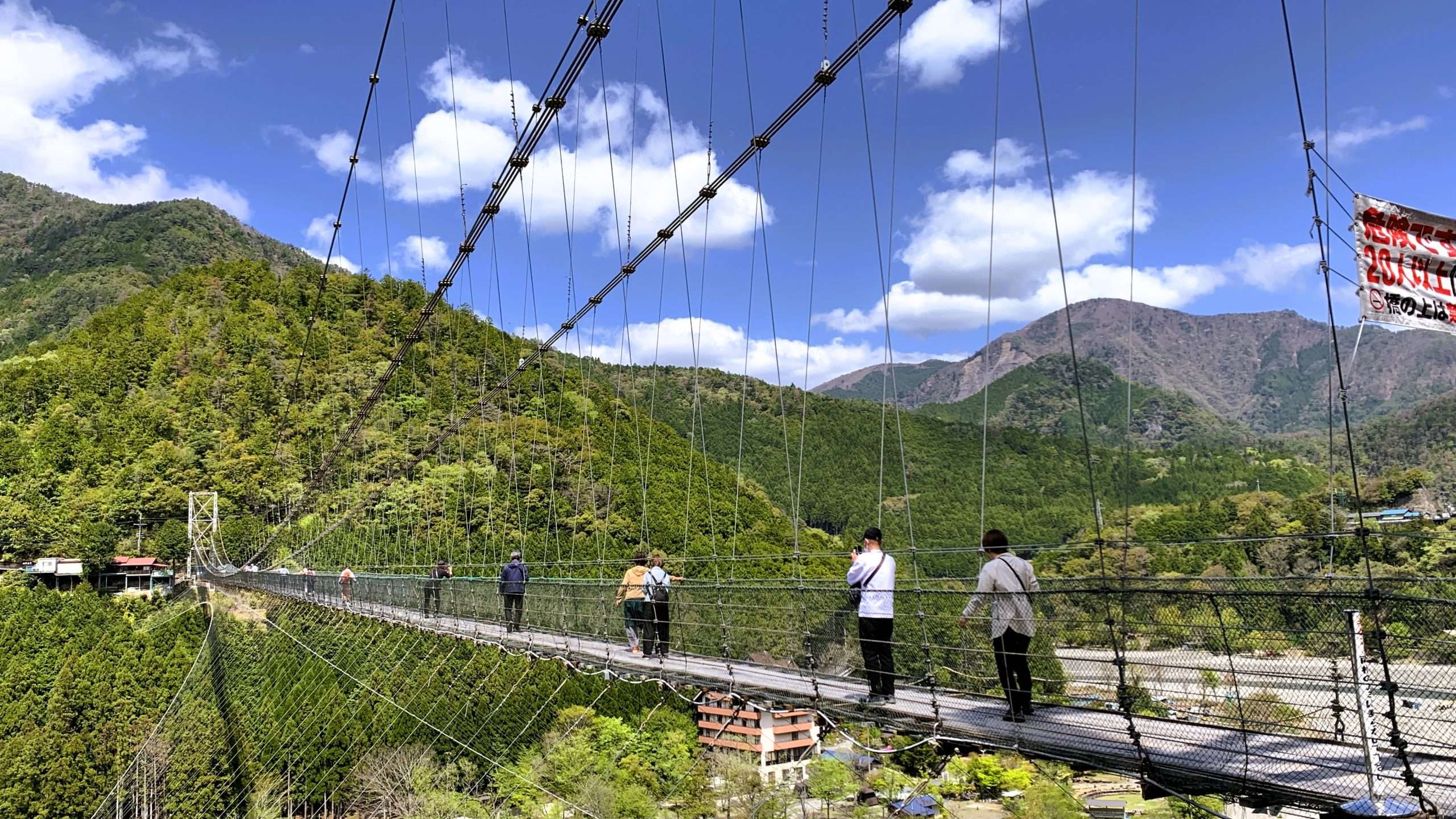 十津川村の観光名所・谷瀬の吊り橋を斜めアングルで撮影。297メートルの長大な吊り橋を渡る人々。周囲の深い緑の山々と晴れた青空に少しの雲が浮かぶ、スリルと自然が共存する風景。