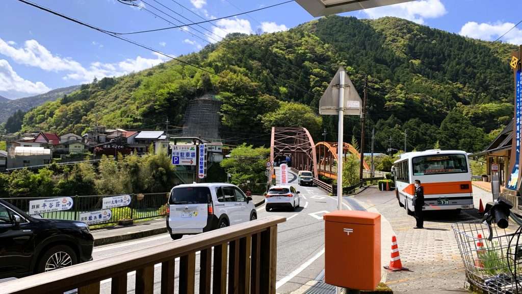 奈良県十津川温泉バスセンター付近を撮影した風景。手前に赤い郵便ポスト、右側バス停に停車するバス、左側道路を走る車、奥には住宅へ続く赤い鉄橋と緑の山々。青い空が印象的な温泉街の景観。