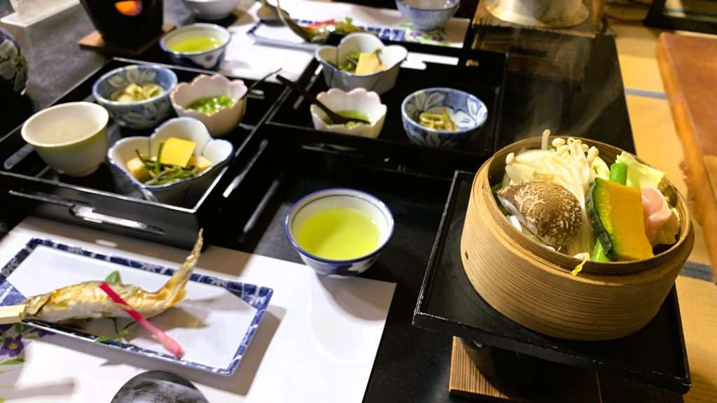 奈良県十津川温泉の民宿やまとやで提供された夕食。右手前に丸いわっぱ容器の蒸し料理があり、カボチャやエノキ、シイタケ、鶏肉、アスパラガスが入っている。アユの塩焼きや山菜料理など地元食材を使った和食が並ぶ温泉宿の食事風景。