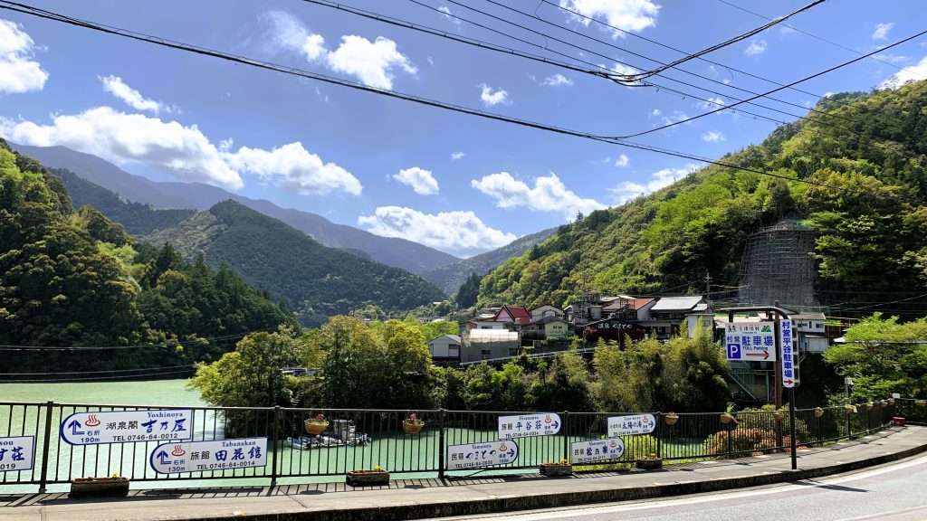 十津川温泉エリアの街並みと自然風景。道路沿いに流れる青緑色の川、温泉街の建物、奥に広がる緑の山々と青空が見える。奈良県十津川村らしい穏やかな景色。