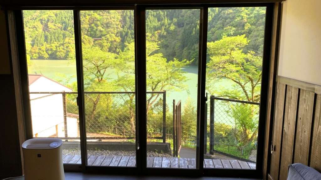 奈良県十津川温泉の民宿やまとや洋室から見える景色。薄緑色に輝く川と周囲を囲む山々、手前の縁側と緑の木々、晴れた日に自然の美しさが広がる温泉宿の風景。
