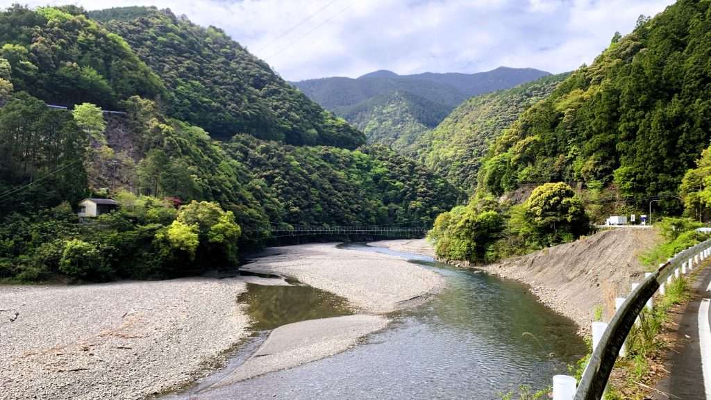十津川村を流れる川と周囲の山々を撮影した風景。晴れた空の下に自然が広がり、遠くに柳本橋の吊り橋が見える奈良県の絶景。