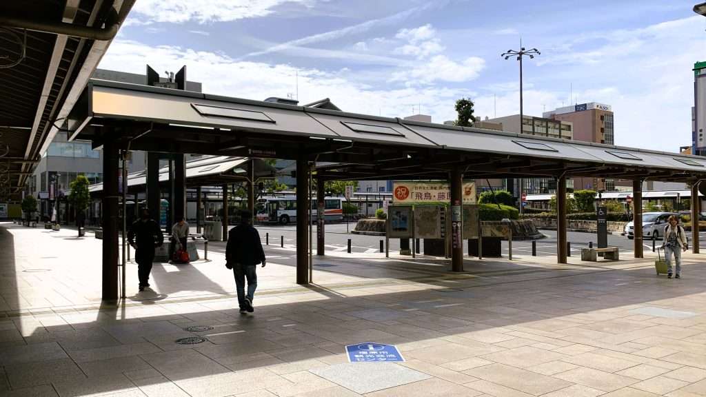 奈良県の近鉄大和八木駅前バスターミナルを撮影した風景。屋根付きバス停に数人の利用客。石畳の広場と整然と並ぶ駅前建物。青空と雲が広がる落ち着いた都市景観。