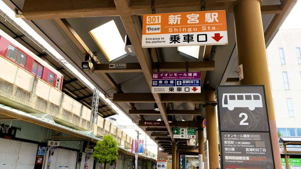奈良県の近鉄大和八木駅前バスターミナル2番のりばを撮影。上部に新宮駅乗車口と書かれたオレンジ色の案内看板、右手にバス停、左側2階には駅ホームと通過中の電車、1階には昔ながらの店舗が並ぶ駅前風景。
