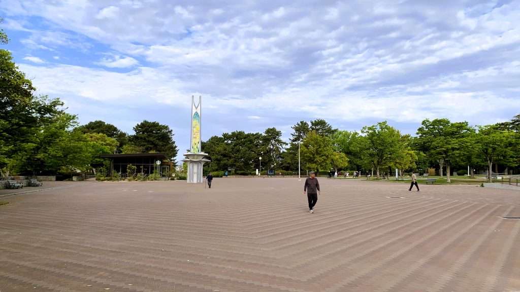 服部緑地西中央広場の様子を撮影。ひまわりデザインのモニュメントと幾何学模様の舗装、奥に見えるスターバックスと緑豊かな樹木、青空と雲の下で人が行き交う大阪の公園風景。