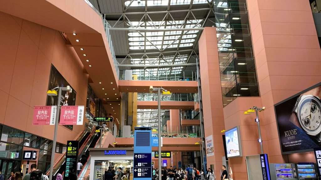 関西国際空港内を見上げる構図で撮影した風景。多くの利用者が行き交う空間。ピンク色の壁が印象的で、上部には通路が架かり、天井から自然光が差し込む開放的な空港施設の内部。