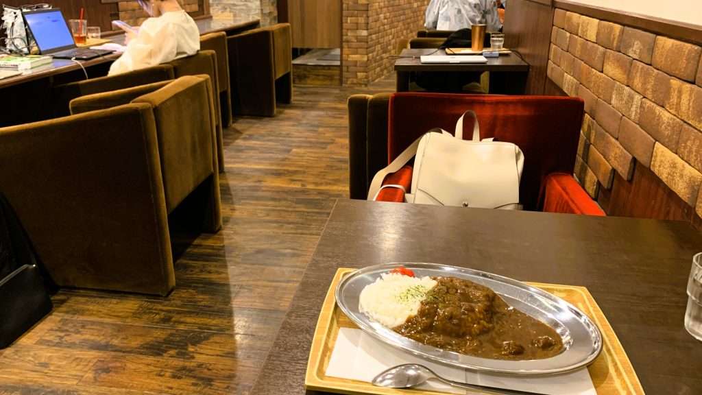 天神橋筋商店街にある横浜珈琲の店内を撮影。木製テーブルに銀の皿で提供されたカレー、レンガ調の壁と木目の床、ビロード調の四角い椅子が並ぶレトロな雰囲気の喫茶店内装。