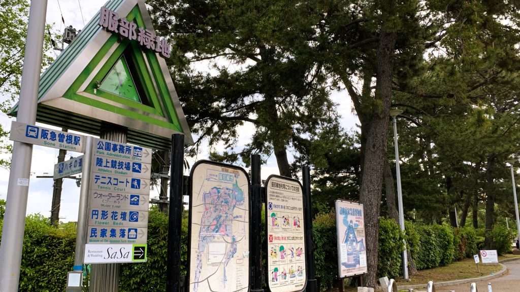 大阪・服部緑地の入口を撮影した風景。「服部緑地」と書かれた木のフォルムの時計と案内板や地図が並ぶエントランス。背後に広がる木々が印象的な公園入口の景観。