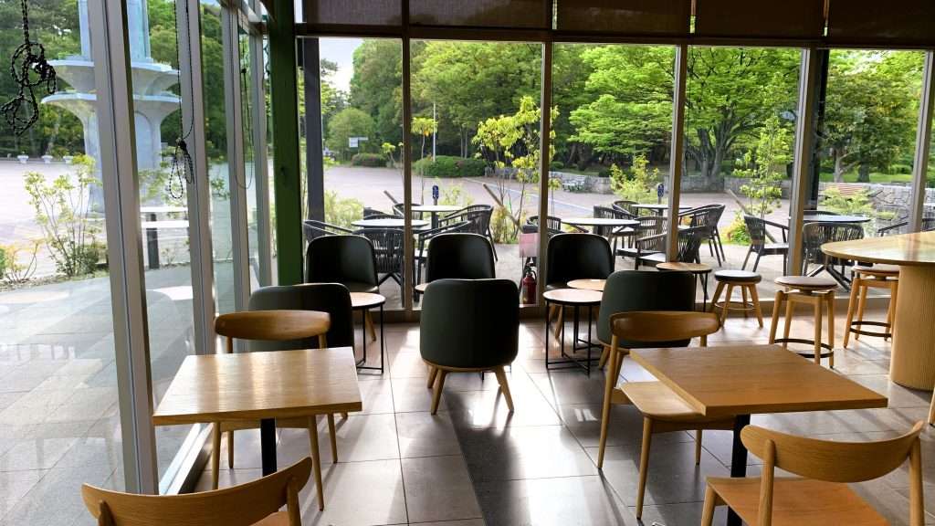 大阪・服部緑地にあるスターバックス店内を撮影した風景。朝早い時間帯で客の姿はなく、一面ガラス張りの窓から公園の緑が見える。木製のテーブルや椅子が配置された落ち着いた洗練されたカフェ空間。