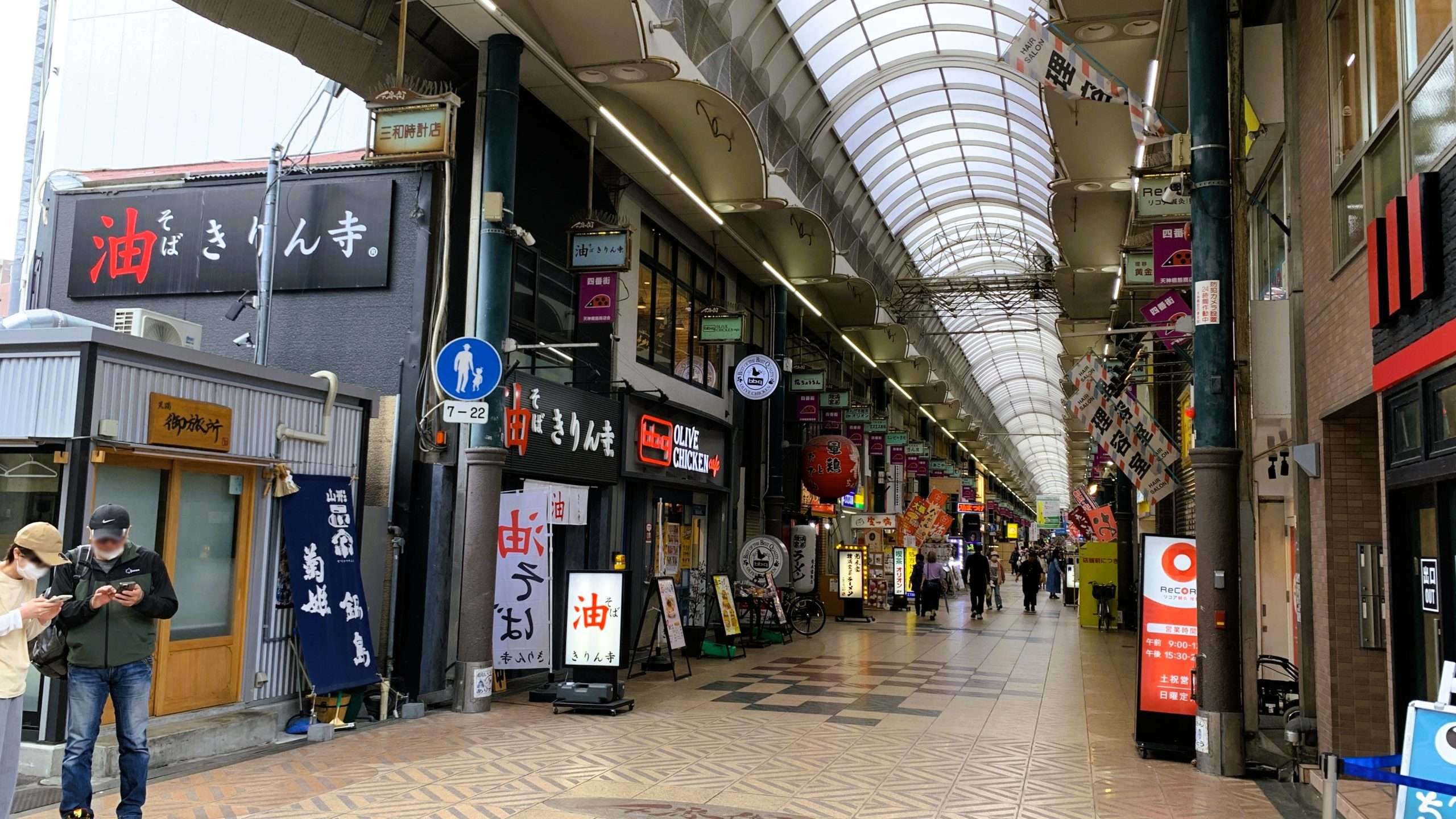 日本一長い商店街として知られる天神橋筋商店街を撮影。左右に多くの店舗が並び、光が差し込む丸アーチ状の天井。左手にスマートフォンで調べ物をする2人、奥にも人通りがある大阪の商店街風景。