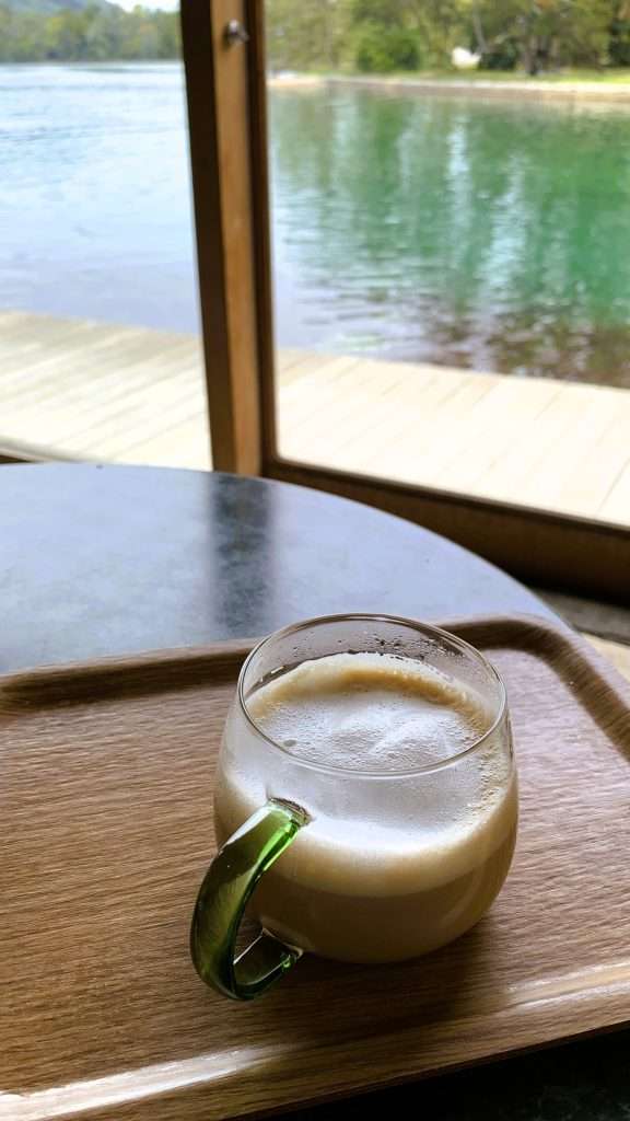 京都・天橋立のカフェ・ドゥ・パン千歳で撮影したカフェ風景。丸いテーブルの上にガラスコップのカプチーノが置かれ、窓の奥には運河と青緑色の水面が見える、落ち着いたカフェ空間。