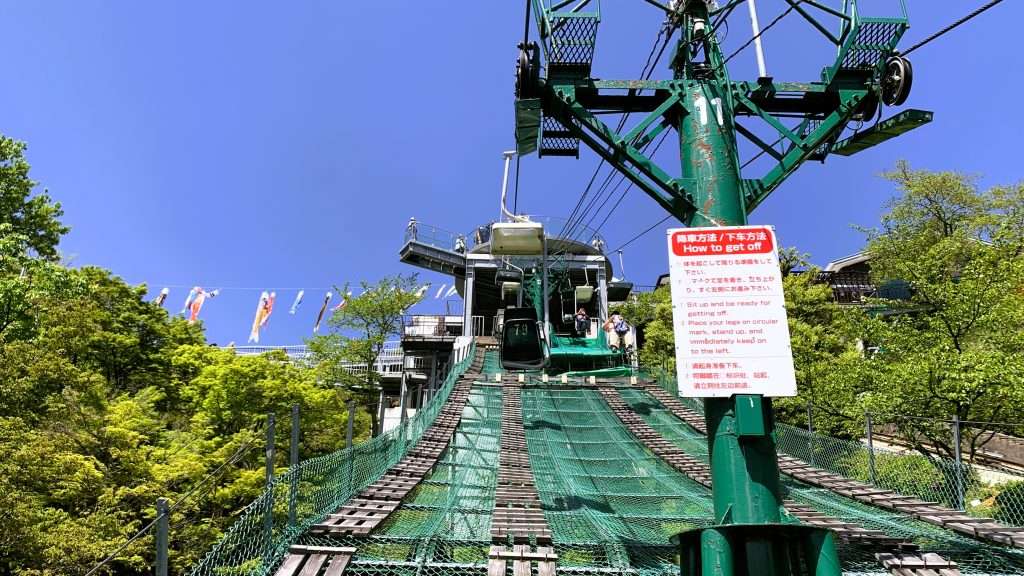 天橋立リフトから見上げるように撮影した景色。府中駅から笠松駅へ向かう途中で山頂駅が見え、右側の支柱に降車案内が掲示され、緑豊かな木々と青空に包まれた京都・天橋立のリフト風景。