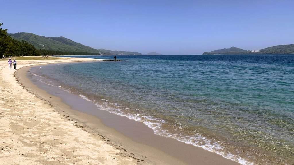 京都・天橋立の海岸から北側を撮影した風景。右側に透き通った青い海、左側に砂浜が広がり、遠くに松並木と山々が見える。快晴の空の下で数人が歩くビーチの自然景観。