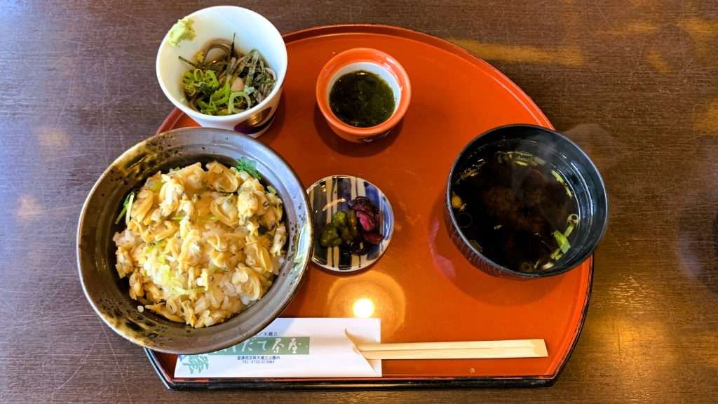 京都・天橋立のはしだて茶屋で提供されたあさり丼を撮影。左側の丼にたくさんのあさりが盛られ、ミニそばや味噌汁、お漬物ともずく酢が付いた定食。赤い半円形プレートと割りばしが並ぶ食事風景。