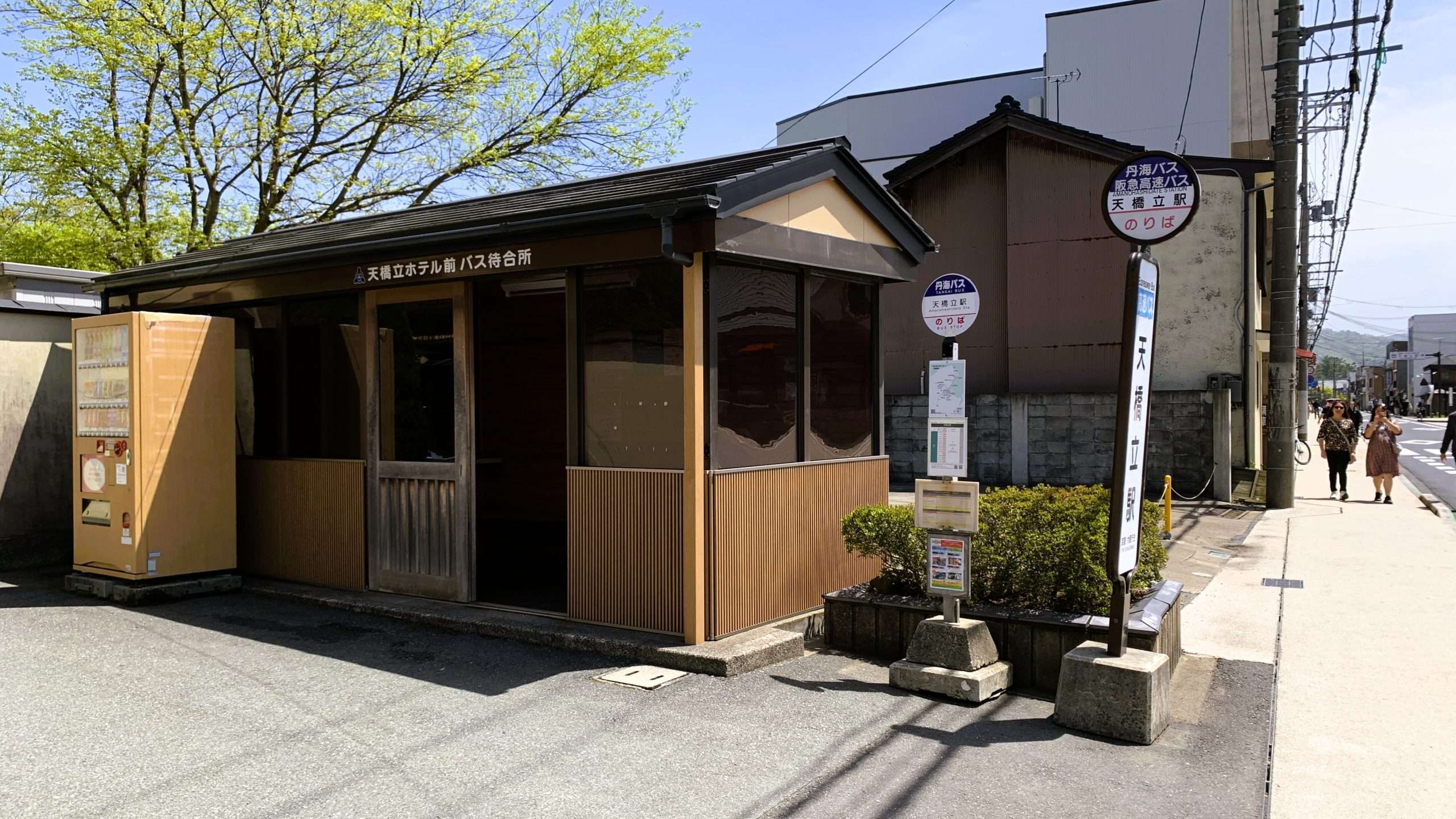 京都・天橋立ホテル前のバス待合所とバス停を撮影した風景。木造の屋根付き待合所の入口に「天橋立ホテル前バス待合所」の看板が掲げられ、右側にバス停標識が並ぶ、青空の下で観光客が歩く天橋立の交通風景。
