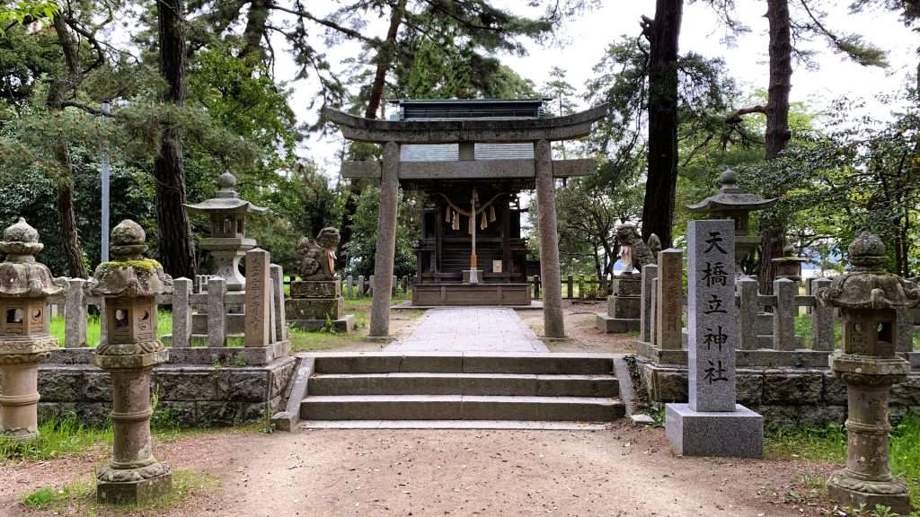 早朝の京都・天橋立神社を正面から撮影した風景。石の鳥居の奥に社殿が建ち、左右に狛犬、手前右側に「天橋立神社」の石碑、参道には石灯籠が並び、松林に囲まれた人影のない静かな神社の景観。