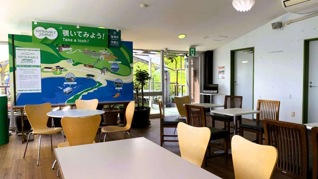 天橋立・笠松公園の2階展望スペースを撮影した風景。白いテーブルと木製チェアが配置された静かな休憩スペース。左側に天橋立の案内イラストパネル、白を基調とした内装と木の床が調和する展望施設。