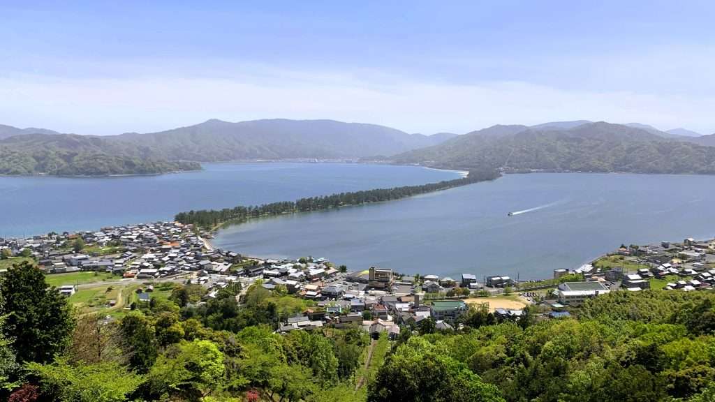 京都・天橋立の笠松公園から撮影した風景。海の上に松並木が一直線に伸びる天橋立の絶景で、青い海と空の下を船が並行して進む姿も見られる。