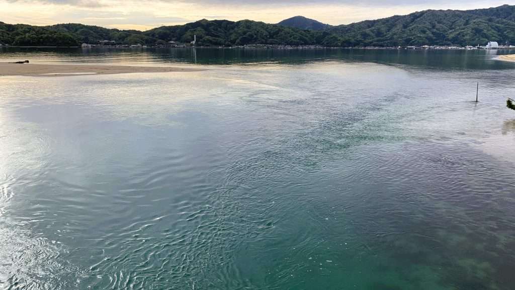 京都・天橋立の海岸を早朝に撮影。海流が交わる場所に波紋が広がり、遠くに山並みと左側の砂浜が見える。曇り空の切れ間から光が差し込む穏やかな海辺の風景。