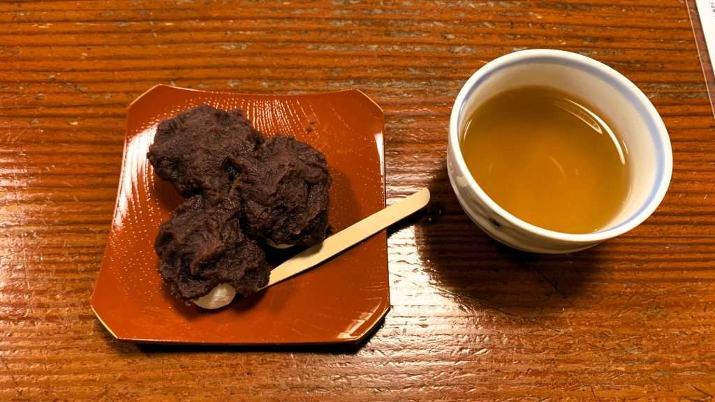 京都・天橋立名物、ちとせ茶屋の知恵の餅を撮影した写真。やわらかい餅にあんこを包んだ和菓子が朱色の皿に3個並び、木製の菓子楊枝とほうじ茶が添えられた天橋立を代表する名物甘味。