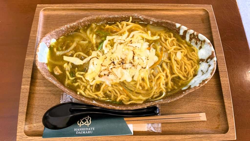 京都・天橋立の橋立大丸本店レストランで提供されたカレー焼きそばを撮影。楕円形の茶色い陶器の器に盛られ、とろみのあるカレースープとチーズが絡むまろやかな焼きそば料理、茶色いテーブルとトレーの上に置かれた食事風景。