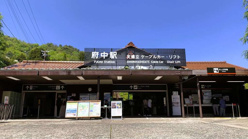 京都・天橋立ケーブルカー・リフトの府中駅を正面から撮影した風景。オレンジ色の瓦屋根を持つ平屋の駅舎に「府中駅 天橋立ケーブルカー・リフト」の案内表示、中央にのりば案内、右側に切符売り場があり、青空と緑が広がる観光施設の入口。