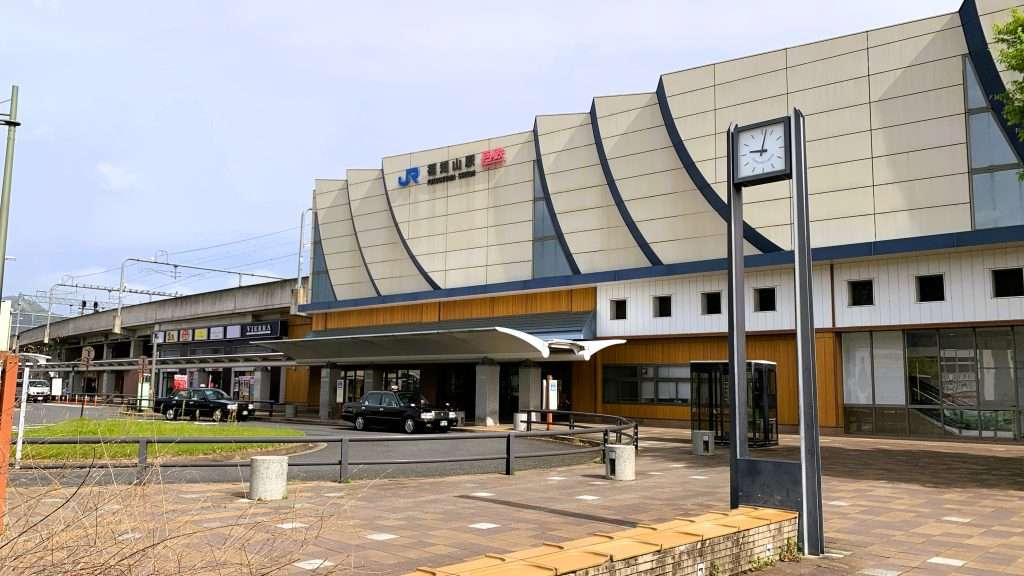京都・福知山駅の外観を撮影した風景。白い壁に青い曲線デザインが施された特徴的な駅舎。1階には木材を使った入口や店舗があり、駅前には歩道、時計、2台のタクシーが見える晴れた日の静かな駅前風景。