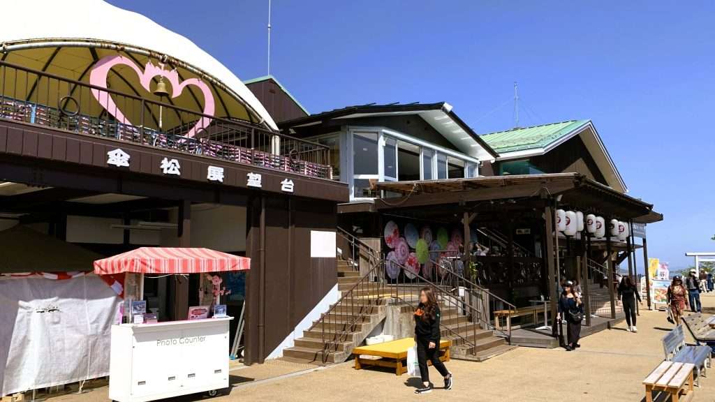 京都・天橋立の笠松公園を撮影した風景。笠松展望台や土産物店が入る二階建ての茶色い建物が建ち、入口上部に提灯が並び、右側には長いベンチと散策する人々が見える観光スポットの景観。