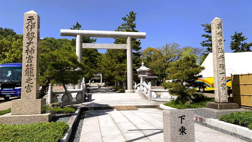 京都・天橋立の籠神社入口を撮影した風景。白い石造りの鳥居と石橋、まっすぐ続く参道。両脇に「元伊勢大神宮籠之宮」と「丹後一宮」の石碑が立ち、青空と緑の木々に囲まれた歴史ある神社の入口。